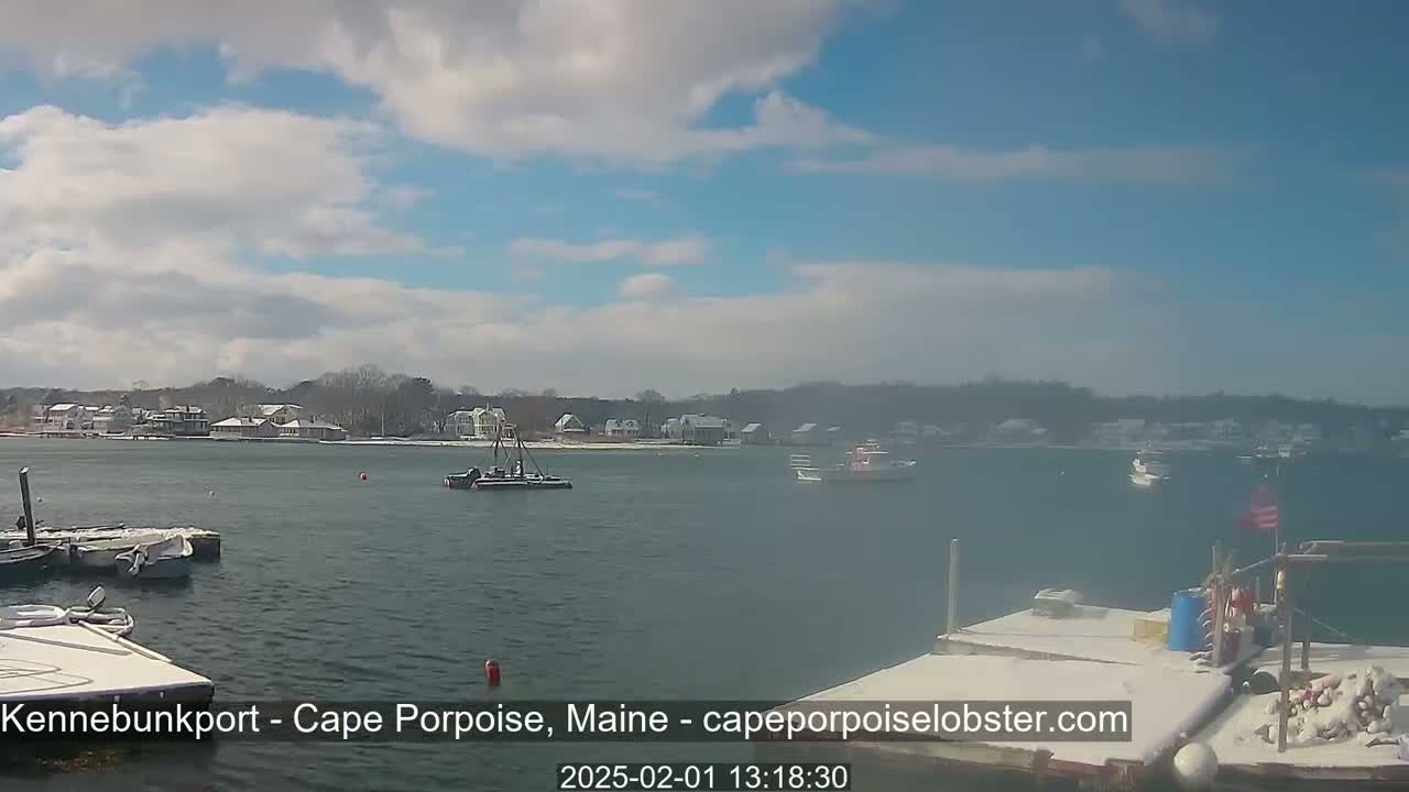A partly cloudy day shows a calm body of water with several boats, a snow-covered shoreline with houses, and a snow-covered dock in the foreground.