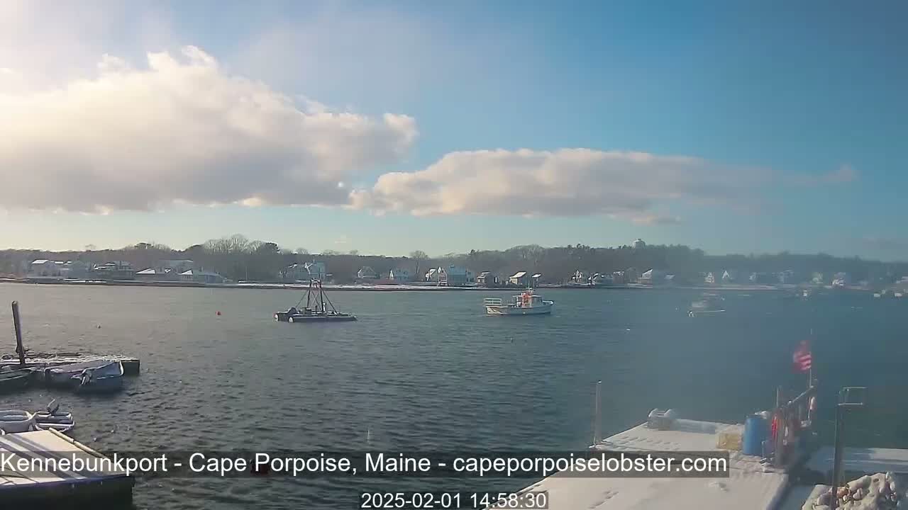 A partly cloudy day shows a calm body of water with several small boats and a snow-covered shoreline with houses in the background.