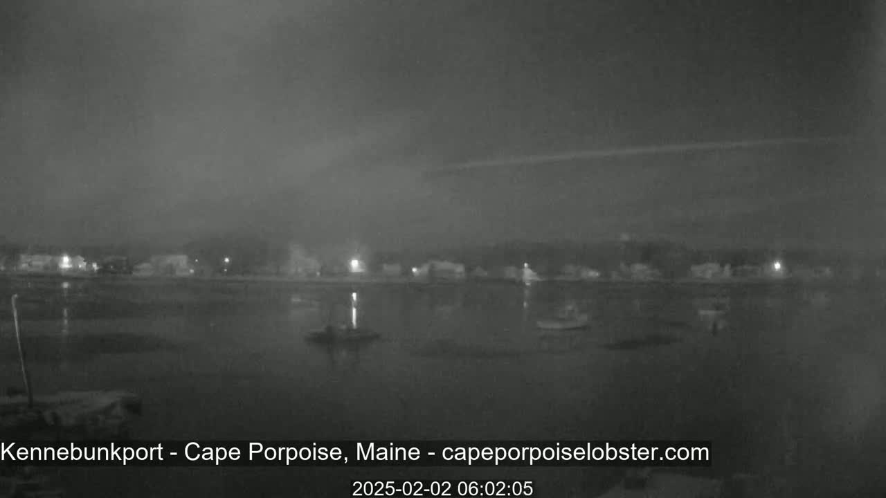 A nighttime, overcast view of a calm body of water with a few boats and a shoreline with buildings visible in the distance.