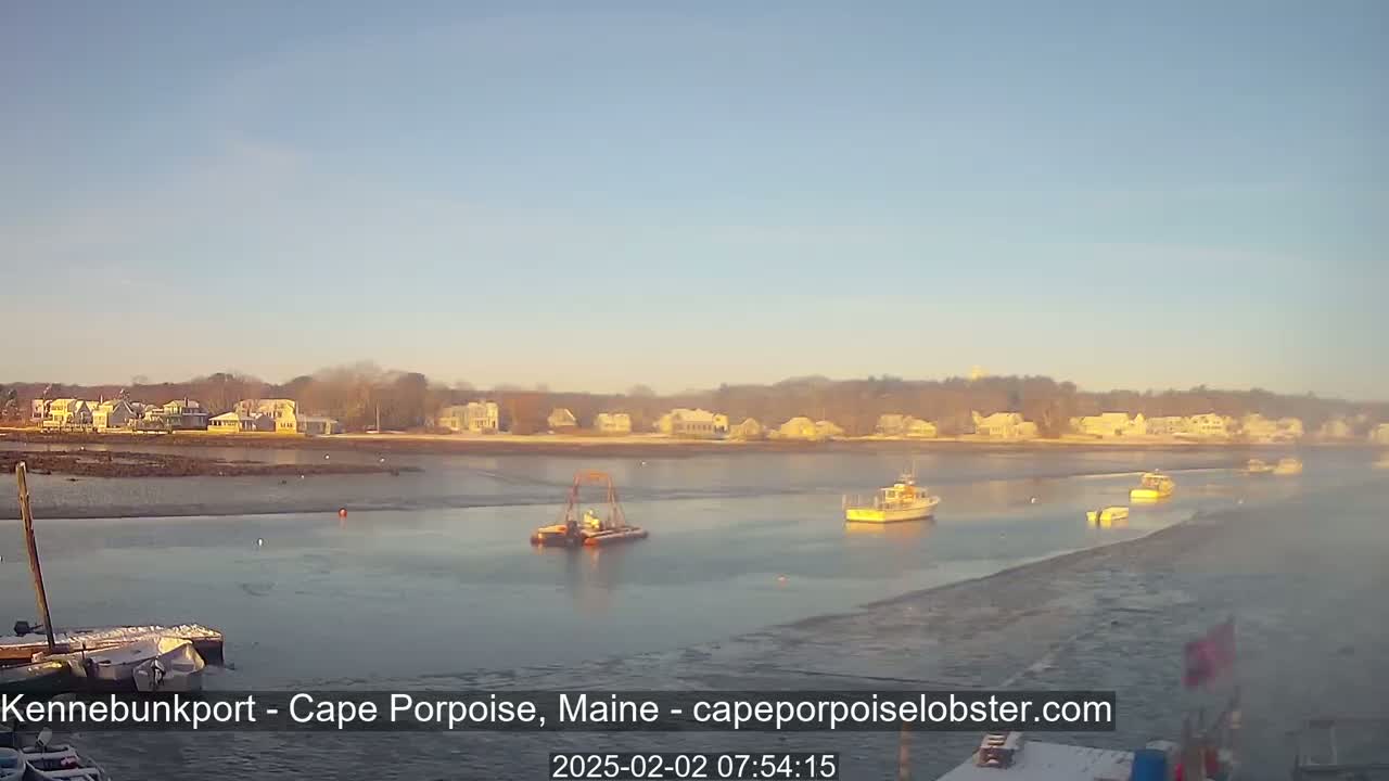 A calm, partly frozen body of water with several boats, some partially frozen docks, and a shoreline of houses under a clear, bright sky.