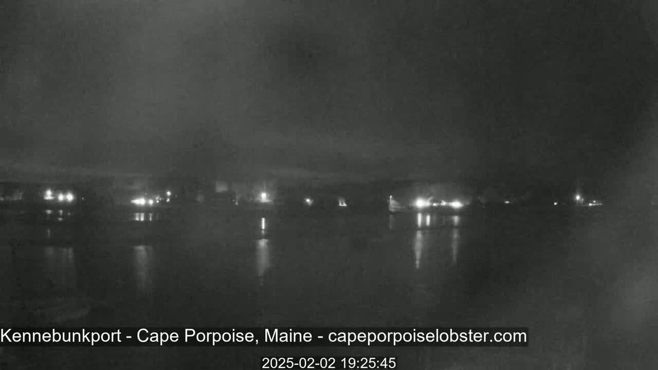 A nighttime view of a calm body of water reflecting the lights of a distant town under a hazy, dark sky.