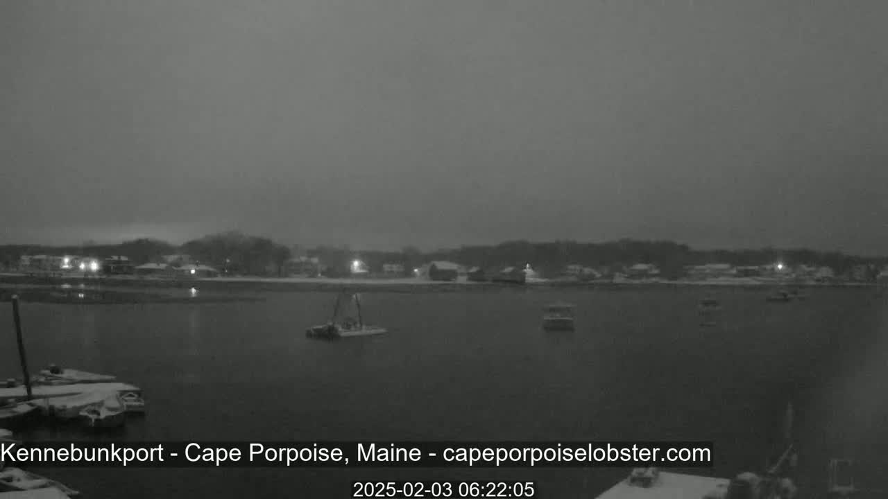 A snow-covered waterfront town is seen at night under a dark, overcast sky, with several boats visible in the calm water.