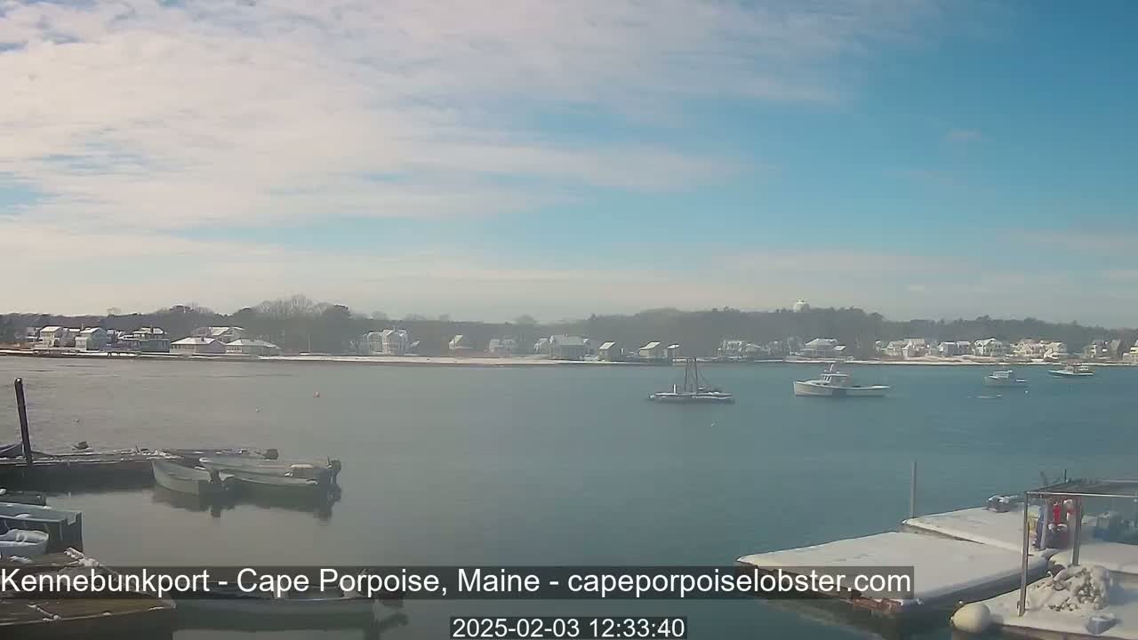 A calm body of water with several boats and a snow-dusted shoreline of houses under a mostly clear, light blue sky.
