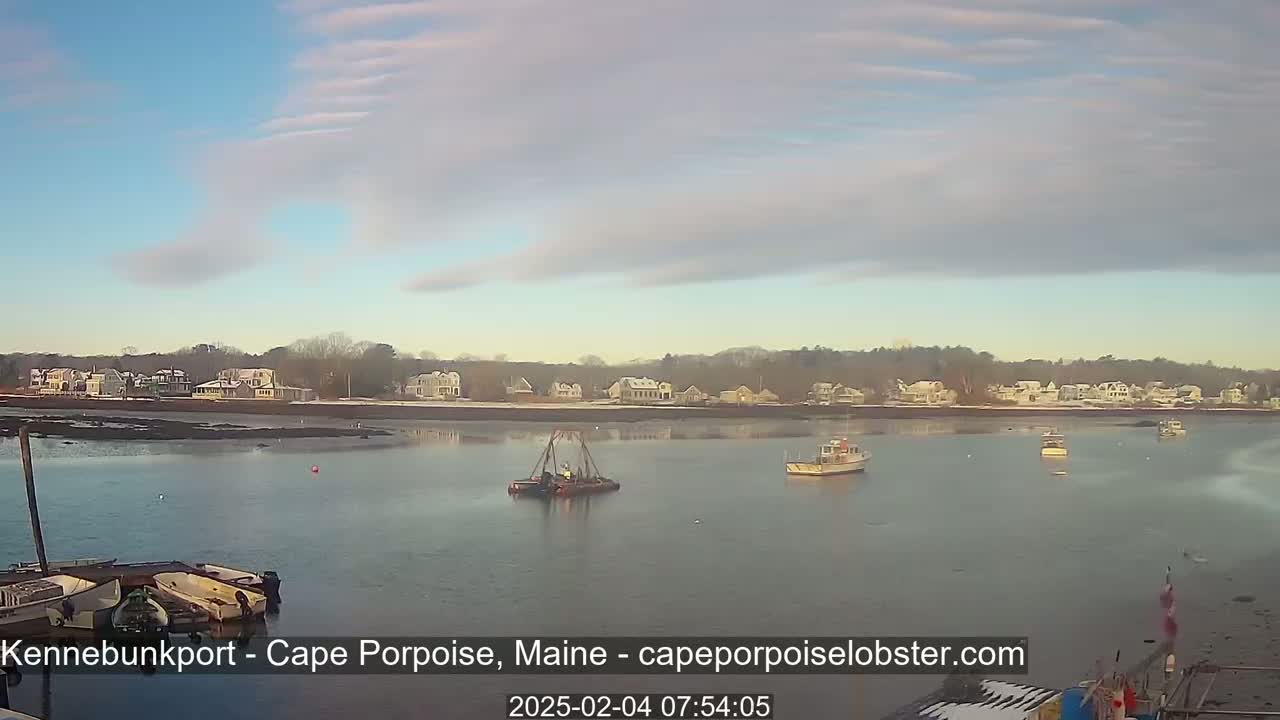 A calm body of water shows several small boats and a larger barge, with a shoreline of houses partially covered in snow under a mostly clear sky.