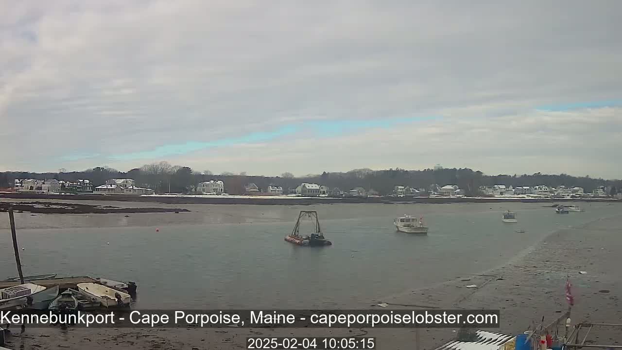 A calm body of water with several small boats, a dredging vessel, and a snow-dusted shoreline of houses under an overcast sky.