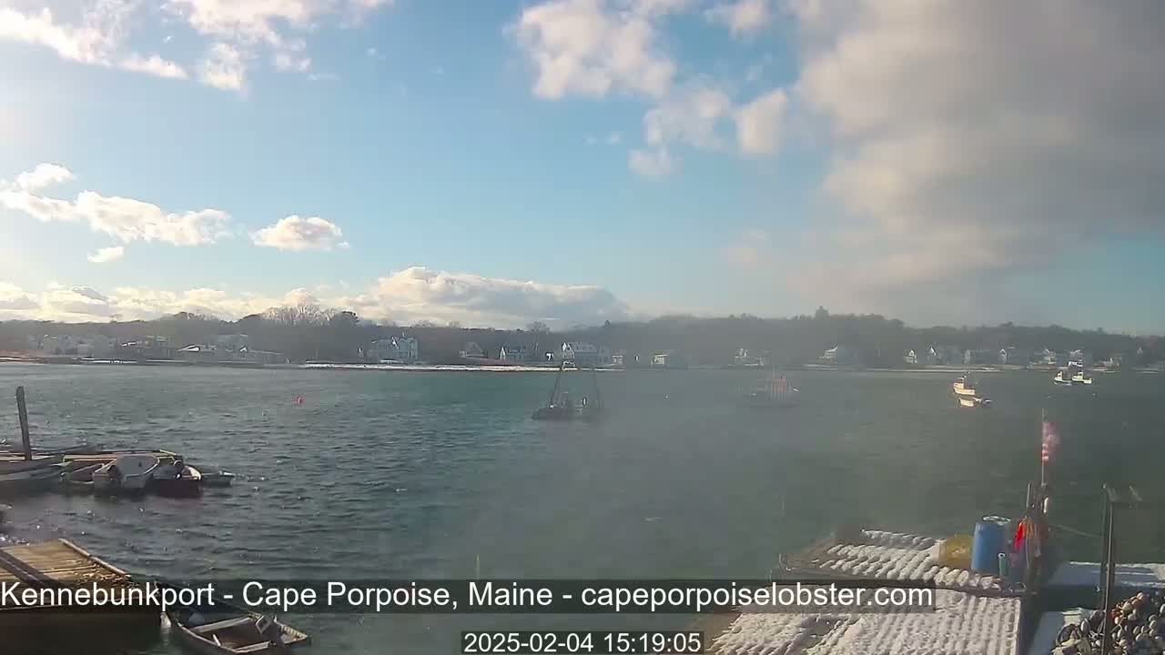 A partly sunny day with some clouds shows a harbor with several small boats and a town in the background, partially covered in snow.