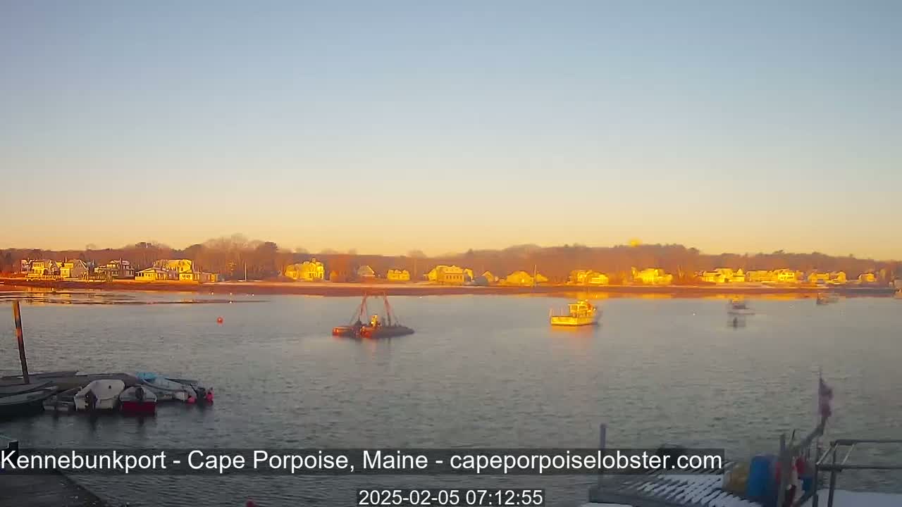 A calm body of water at sunrise shows several boats, including one small barge with a crane, and a line of houses across the water.