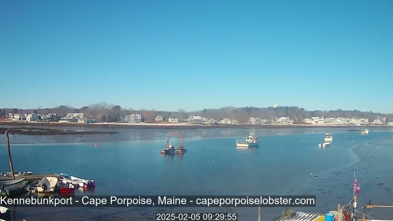 A calm, sunny day shows a harbor with several boats, some docked and others in the water, and houses along the shoreline, partially covered in snow.