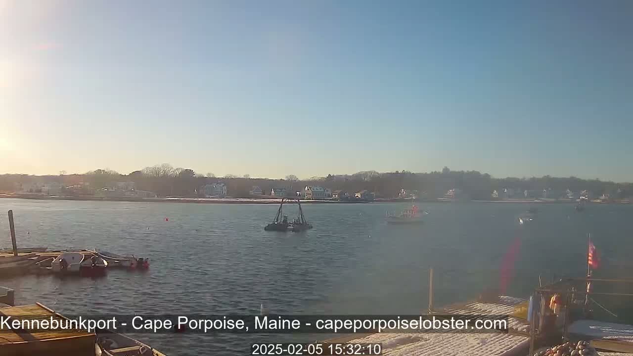 A calm body of water shows several small boats and a structure in the water, with a distant shoreline of houses and trees under a clear, sunny sky.