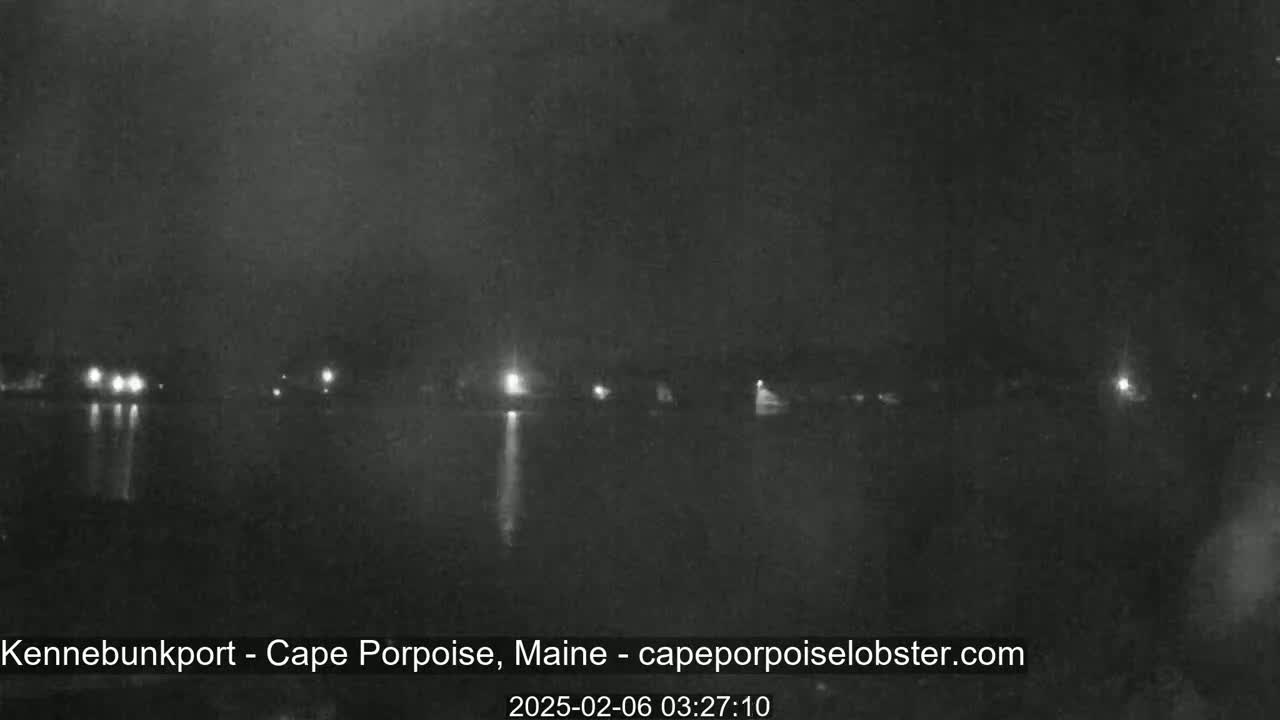 A dark nighttime scene shows a body of still water reflecting a few distant lights on the opposite shore under a cloudy sky.