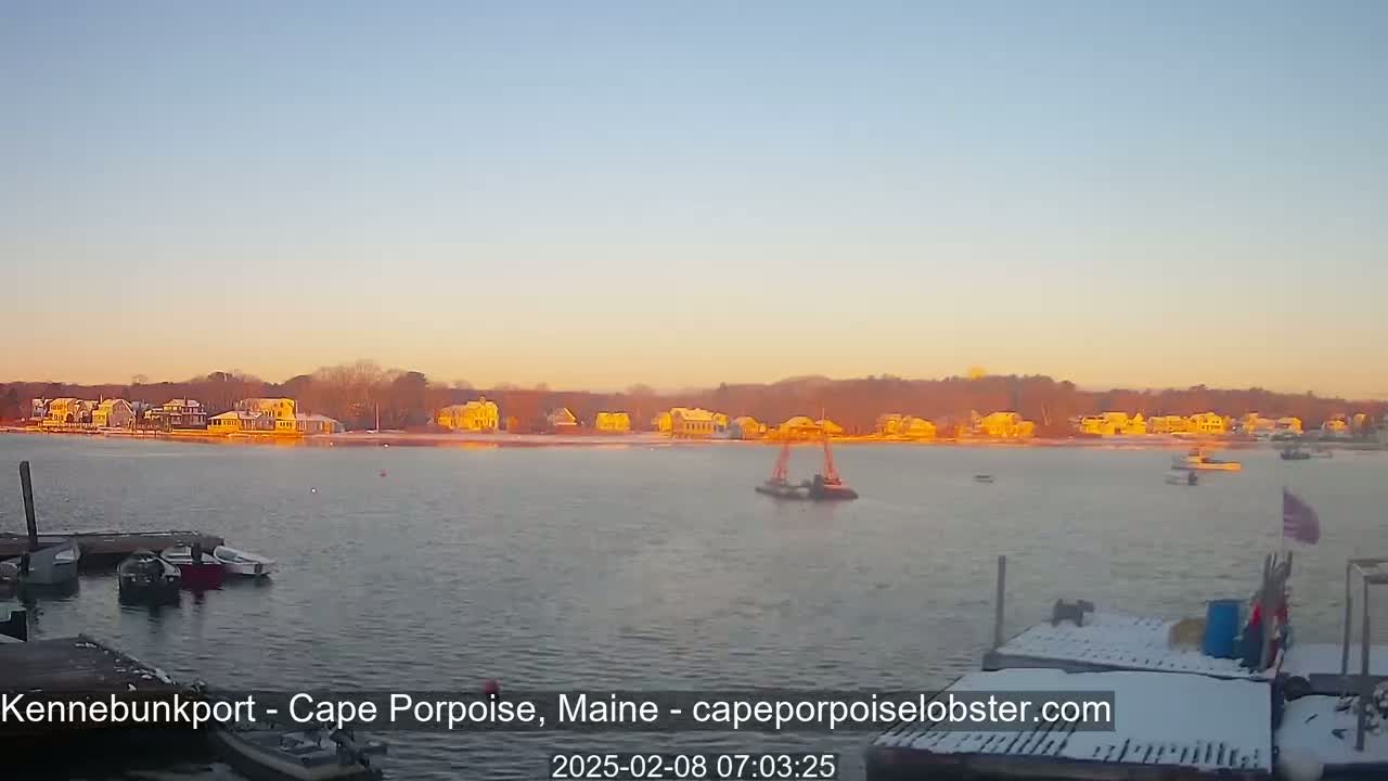 A calm body of water with boats docked at a snow-dusted pier, facing a shoreline of houses bathed in the warm glow of sunrise.