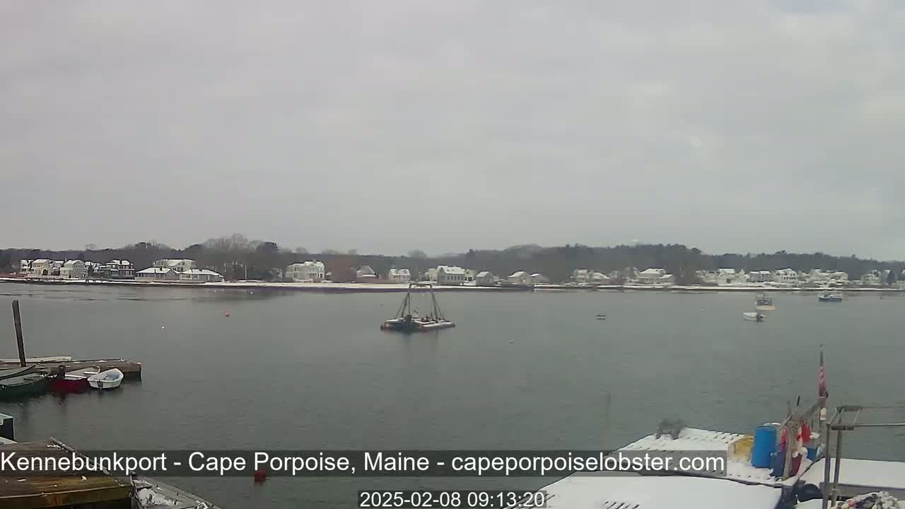 A calm body of water with a few boats, a snow-covered shoreline with houses, and a cloudy sky.