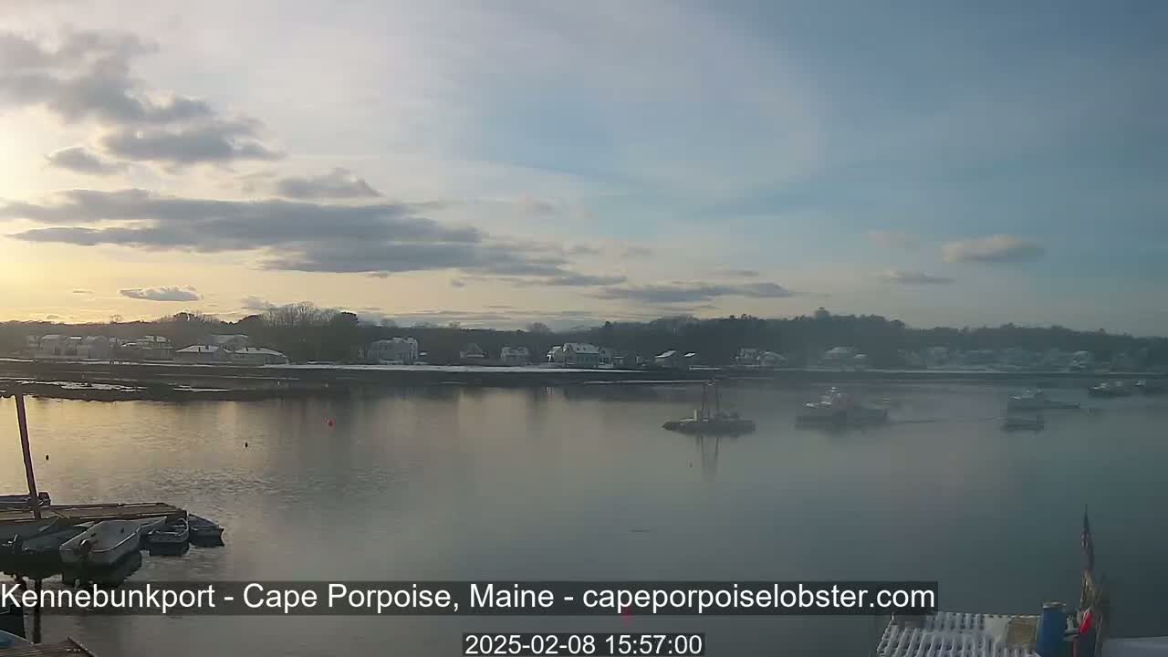 A calm body of water with several boats is seen at sunset, under a partly cloudy sky, with a snow-covered shoreline in the background.