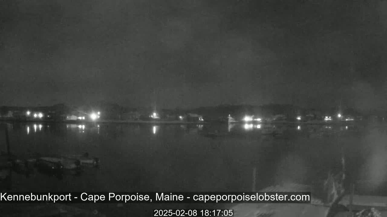 A nighttime view of a calm body of water reflecting the lights of a town across the shore under a dark, cloudy sky.