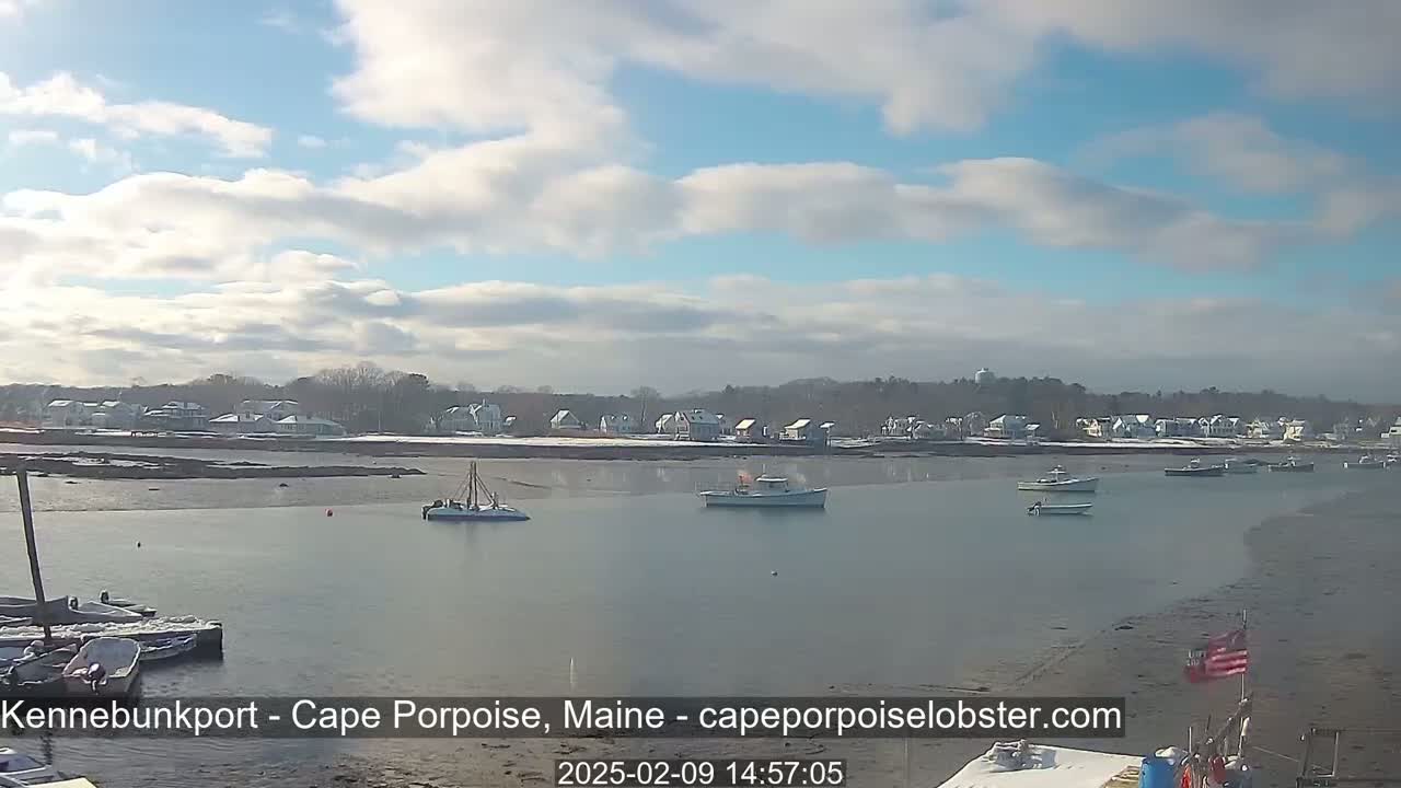 A partly cloudy day reveals a calm harbor with several boats and a snow-dusted shoreline featuring houses.