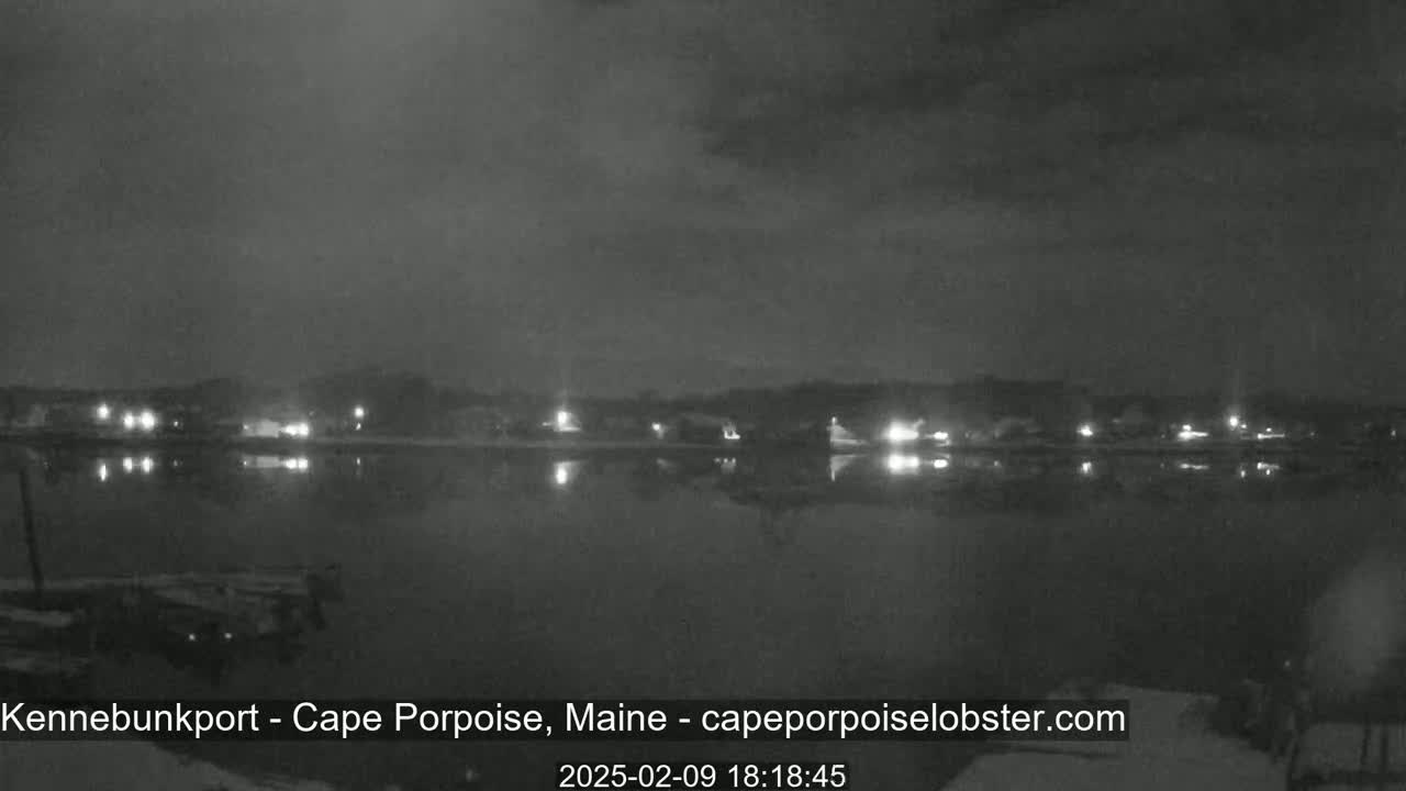 A nighttime view of a calm body of water reflecting the lights of a town across the shore under a cloudy sky.