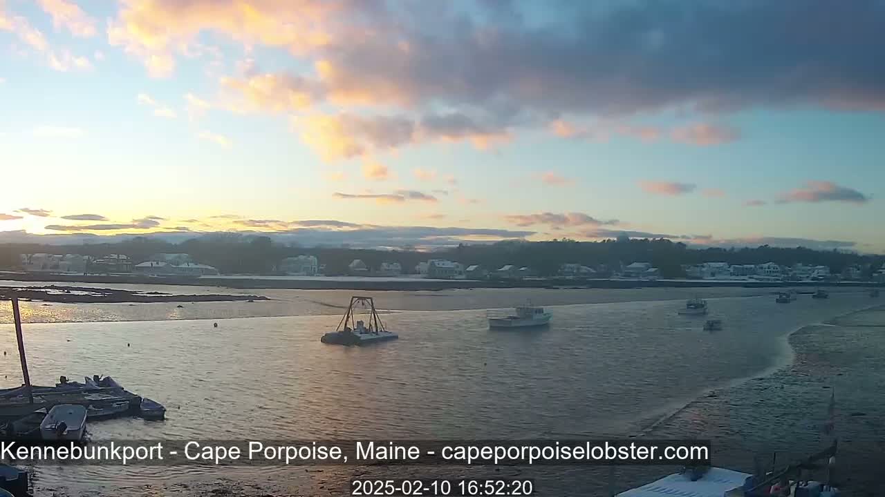 A calm body of water with several small boats is seen at sunset, with a snow-covered town in the background.