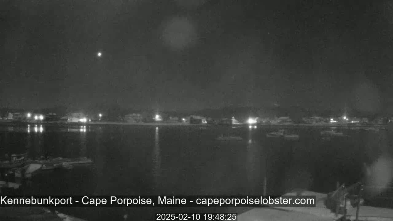 A nighttime view of a calm body of water with a town's lights reflecting on the surface under a dark, clear sky.