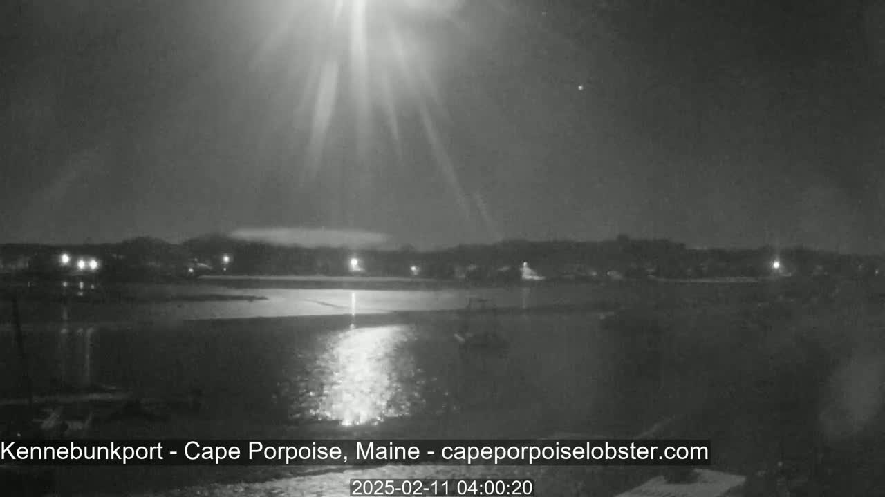 A nighttime view of a calm body of water reflecting bright lights from across the shore under a dark sky with bright beams of light emanating from an unseen source.