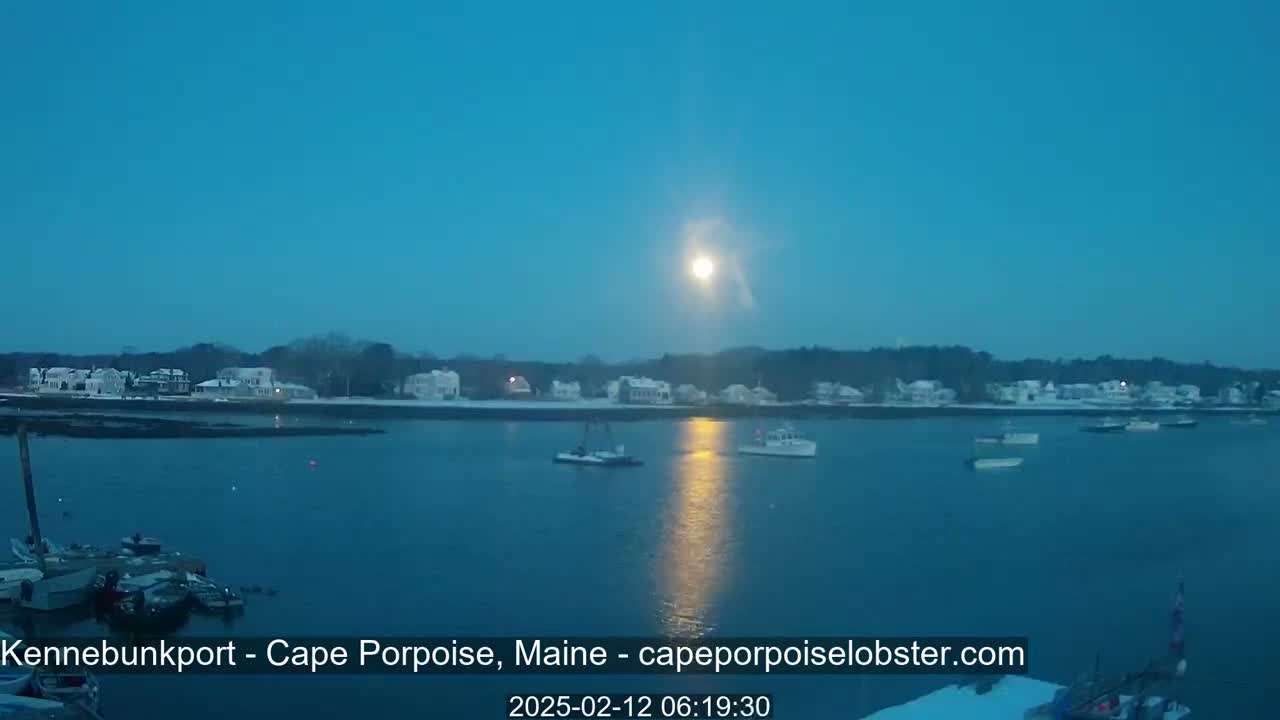 A bright moon illuminates a calm body of water with several boats, a snow-covered shoreline with houses, and a twilight sky.