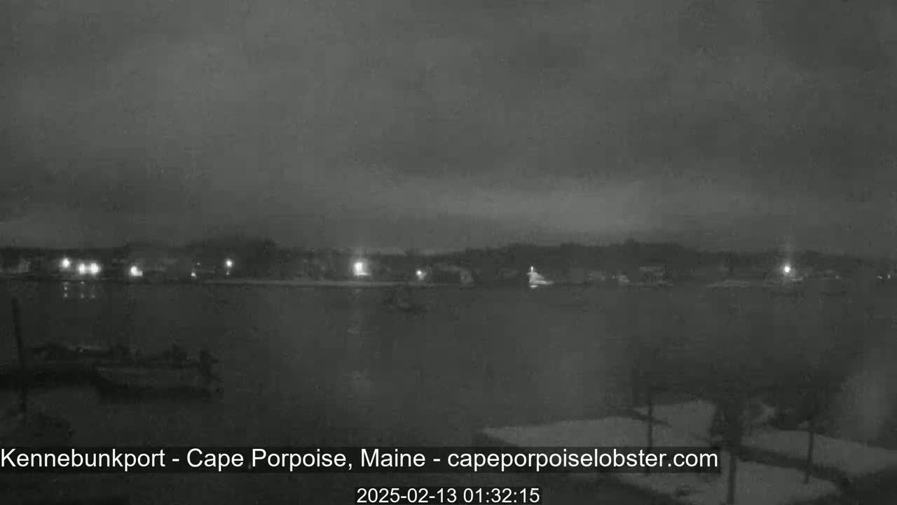 A nighttime, low-light view of a calm body of water with a shoreline showing sparsely lit buildings under an overcast sky.