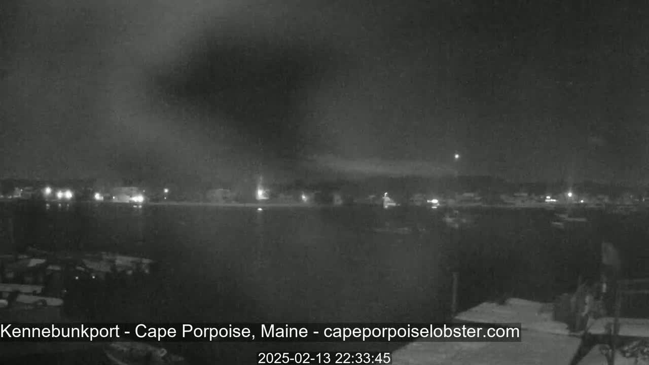 A nighttime long shot shows a dark body of water with dimly lit buildings and vehicles in the distance under a cloudy sky.