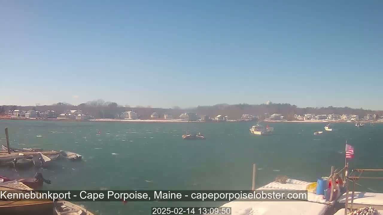 A windy, sunny day shows a harbor with several boats and a snow-dusted shoreline of houses in the distance.
