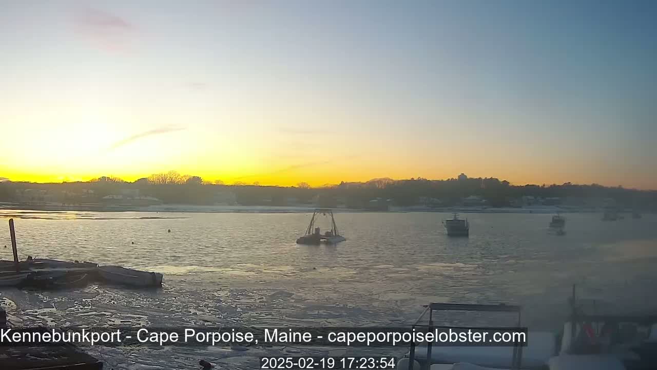 A partially frozen body of water at sunset shows several boats, some docked and others afloat, with a snow-covered shoreline in the background.