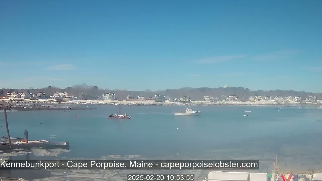 A calm, clear day reveals a coastal scene with a few boats in the water, a snow-dusted shoreline dotted with houses, and some ice along the edges of the water.