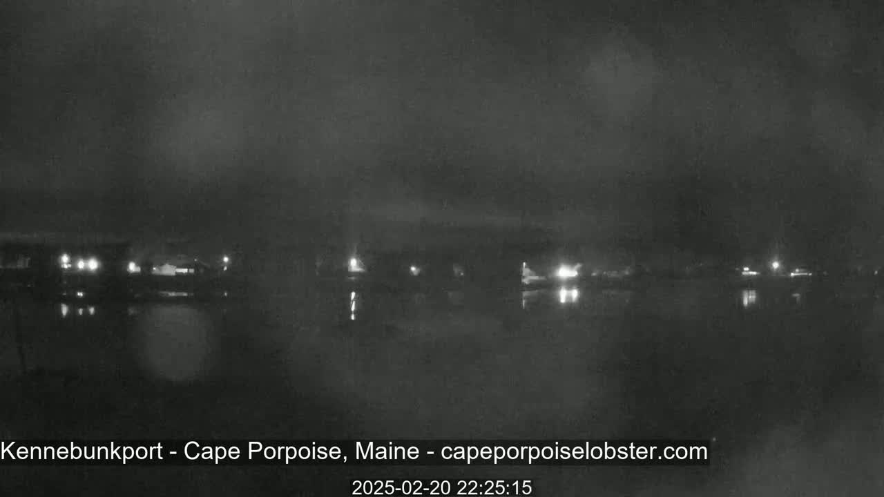 A nighttime, low-light view of a calm body of water reflecting dim lights from buildings across the shore under a dark, possibly overcast sky.