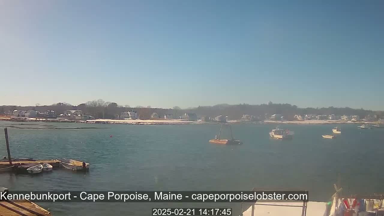 A sunny day shows several boats moored in a harbor with a snow-dusted shoreline in the background.