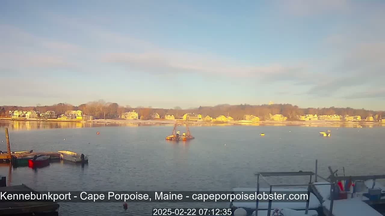 A calm body of water with several boats at a dock,  a dredge in the middle, and a shoreline of houses covered in snow under a clear, slightly hazy sky.