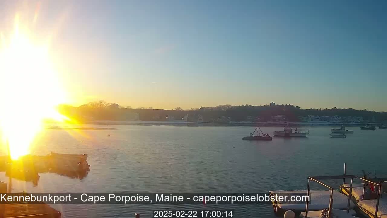 A bright sunset over a calm body of water with several boats, a snow-covered shoreline, and a few buildings in the distance.