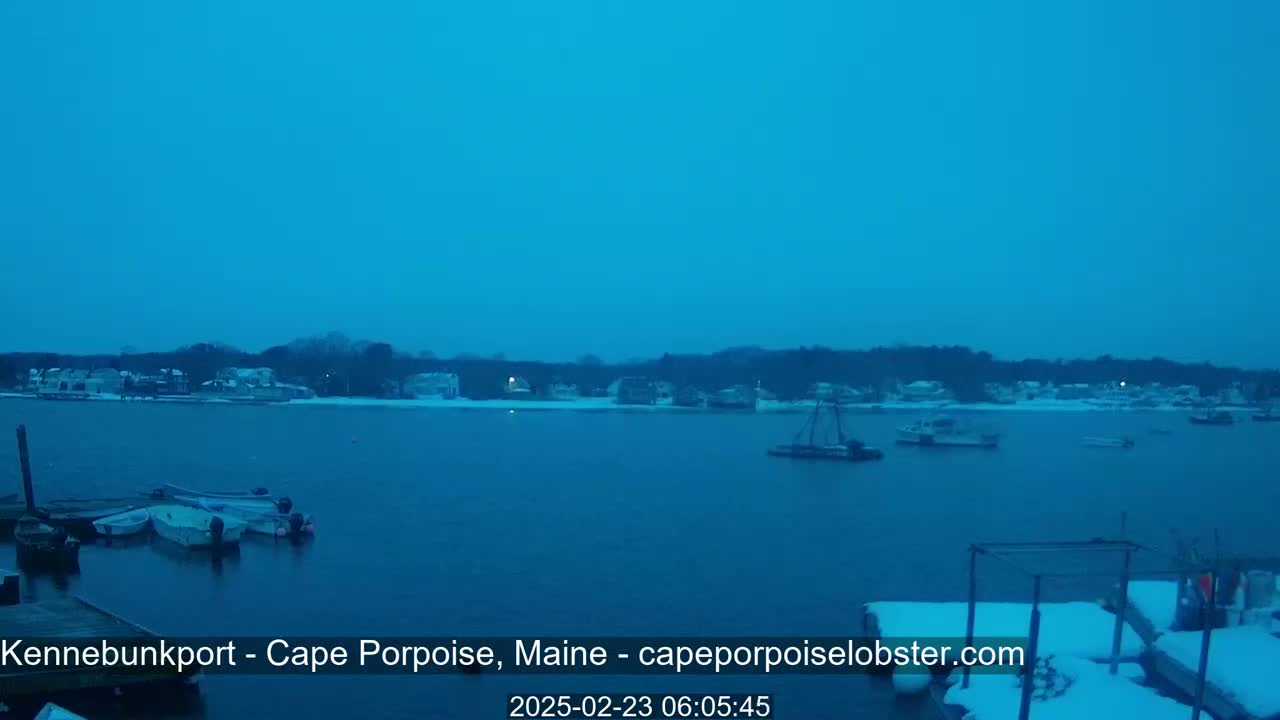 A snow-covered waterfront town is visible across a calm, dark blue body of water, where several boats are moored near a dock.