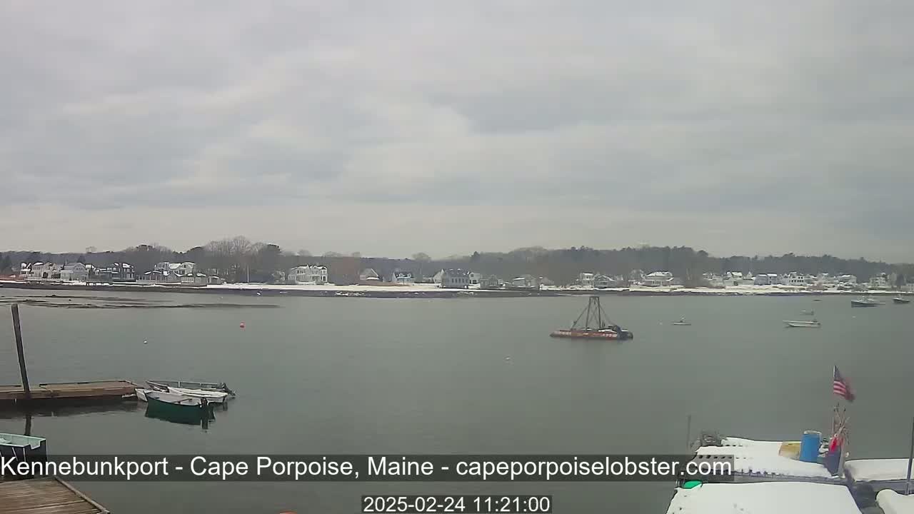 A calm body of water with docked boats and a small vessel in the distance, overlooks a snow-dusted shoreline with houses under an overcast sky.