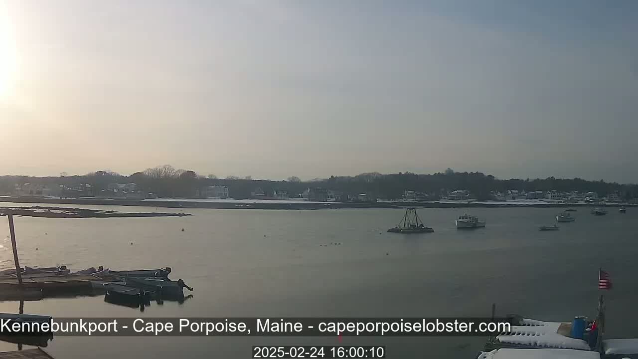 A calm body of water with several small boats, a few larger vessels, and a shoreline of snow-covered buildings under a hazy, bright sky.