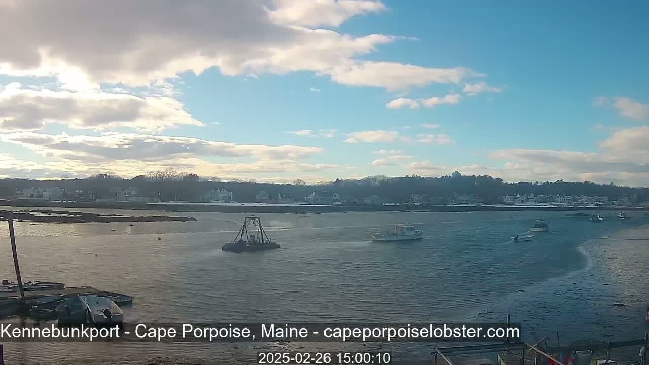 A partly cloudy day shows several small boats on calm water near a shoreline with houses, some of which have snow on the ground.