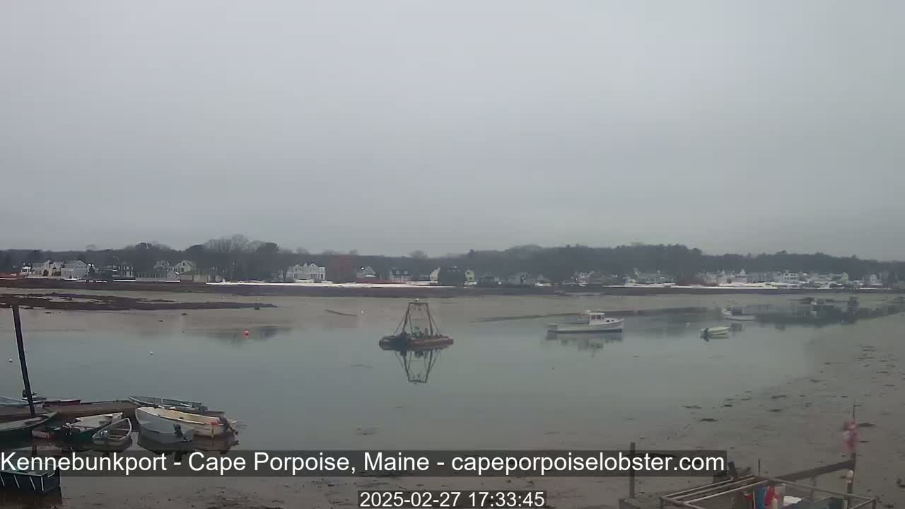 A calm, overcast day reveals a coastal scene with several boats in a harbor, a small dredging vessel, and a shoreline with houses in the distance.