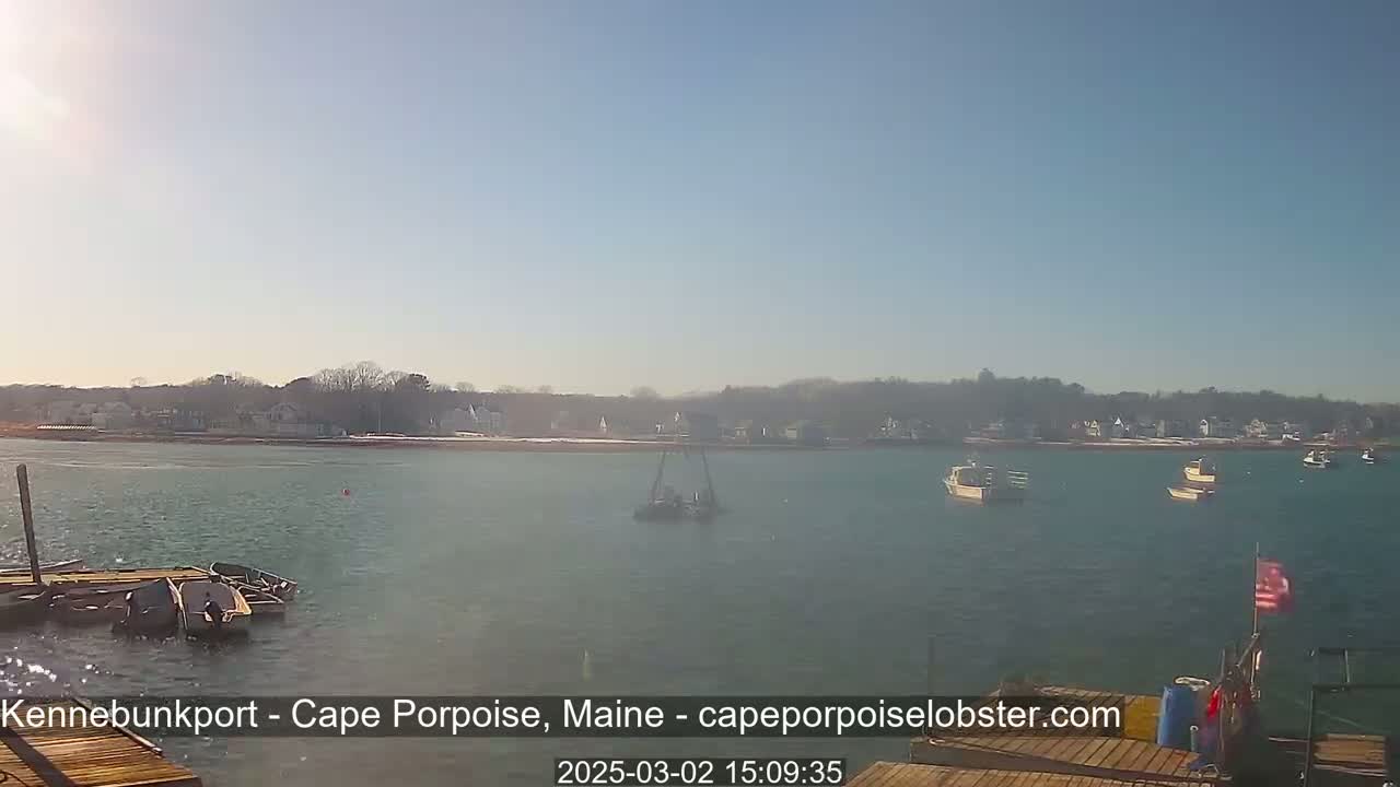 A sunny day shows several small boats on calm water near a shoreline with houses and trees, some snow visible on the ground.