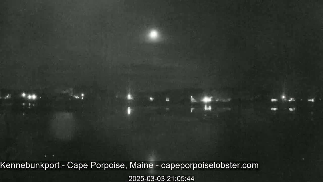 A nighttime, low-light view of a calm body of water reflecting the lights of a distant town under a bright moon.