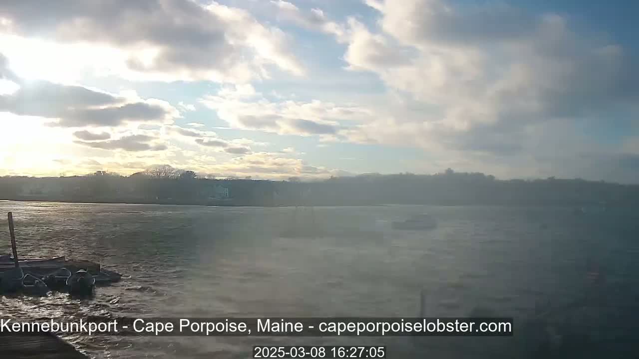 A partly cloudy sky overlooks a choppy body of water with a distant shoreline and several small boats.