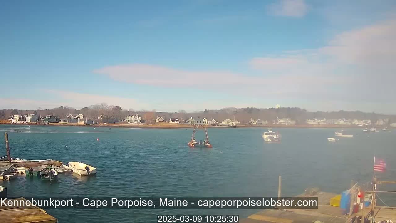 A calm body of water with several boats, a small town in the background, and a partly cloudy sky.