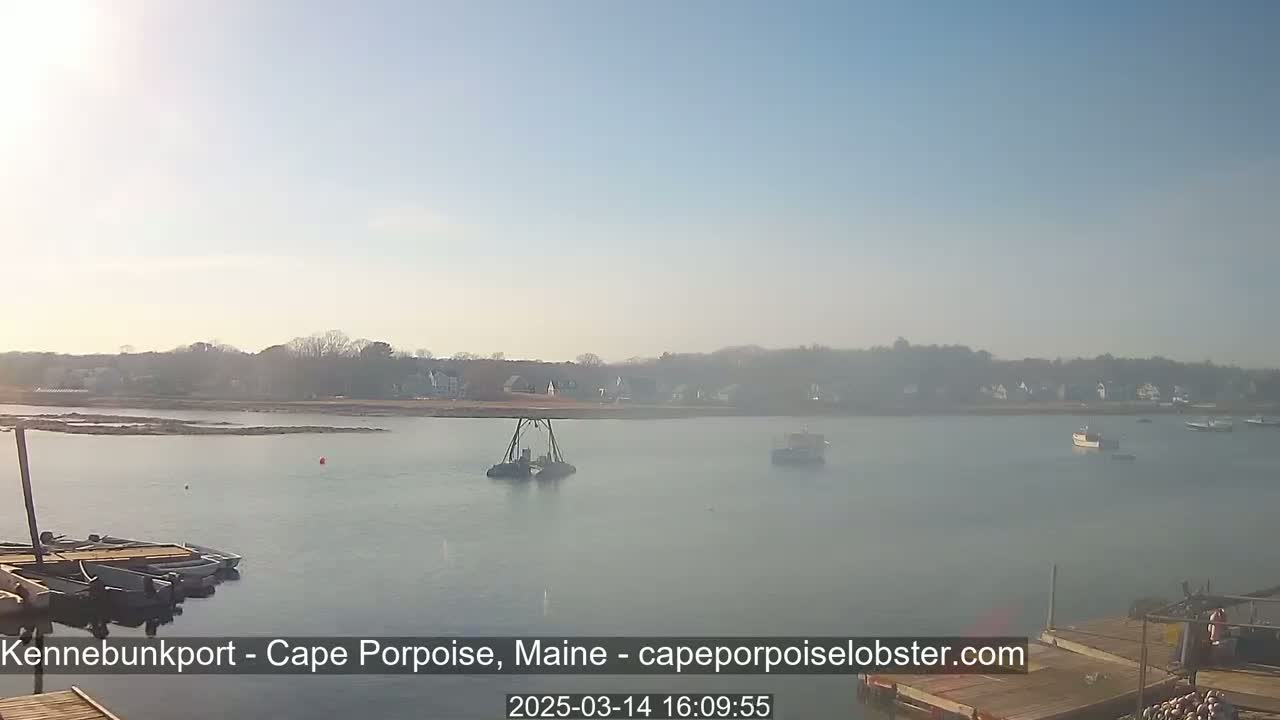 A hazy, sunny day shows a calm body of water with several boats, a small dock in the foreground, and a shoreline with houses and trees in the distance.