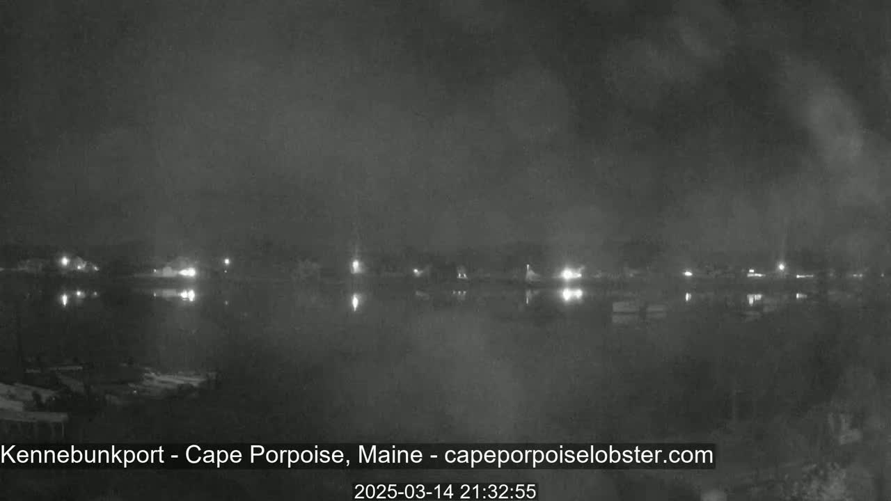 A nighttime, low-light view of a calm body of water reflecting the lights of a distant town under a cloudy sky.