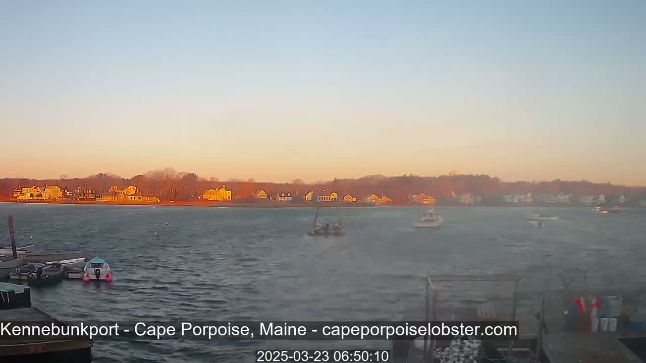 A calm body of water with several small boats and a shoreline of houses under a pale orange sunrise.