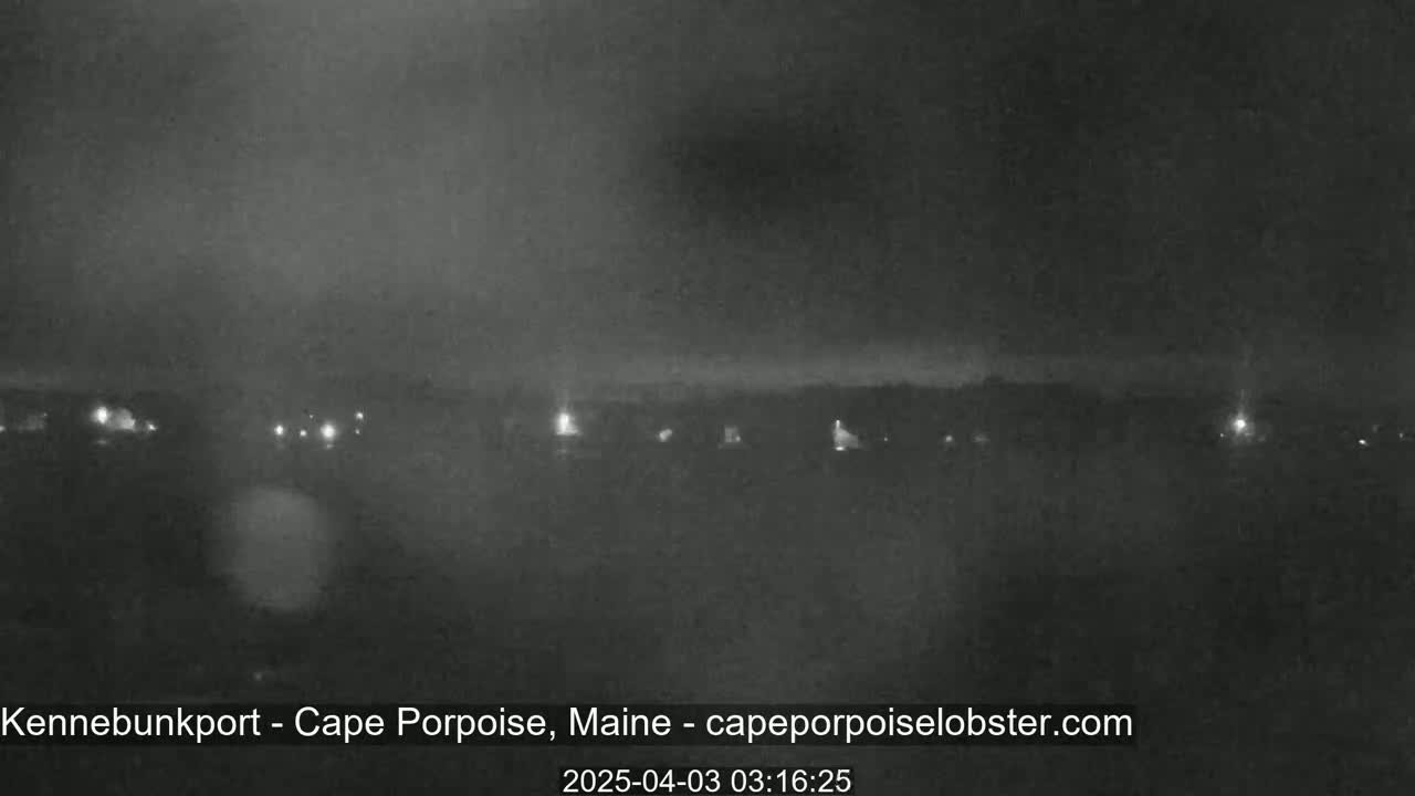 A nighttime long shot shows a dimly lit coastline with scattered lights under a dark, overcast sky.