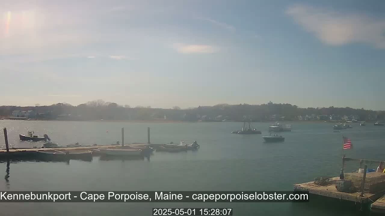 Several boats are moored at a dock in calm water, with additional boats visible further out in the bay under a mostly sunny sky.