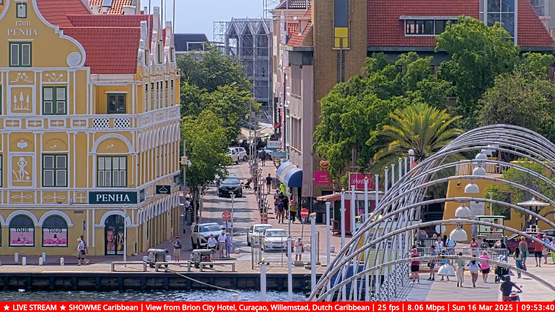 A sunny waterfront street scene shows colorful buildings, pedestrians, cars, and a modern metal walkway arching over a paved walkway.