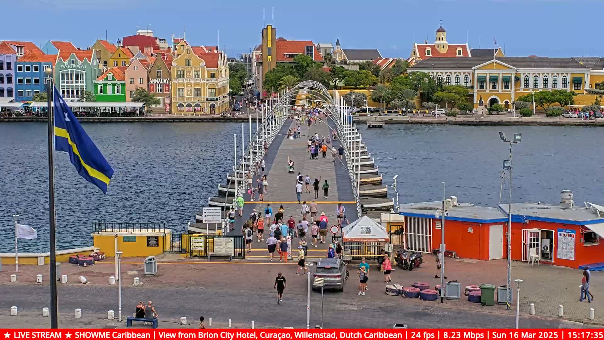 A pontoon bridge with many pedestrians crosses a waterway in front of colorful buildings under a sunny sky.