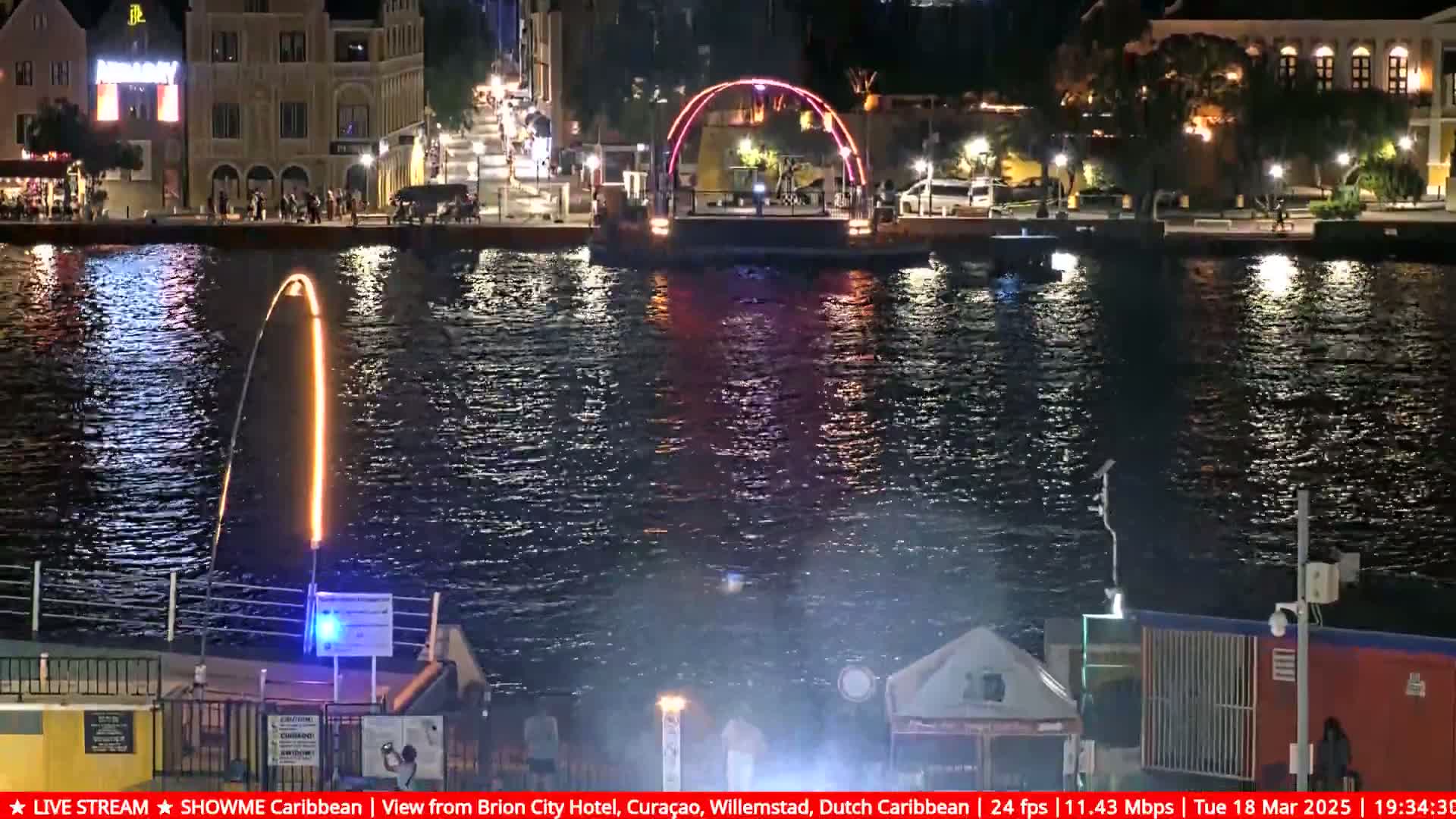 A nighttime waterfront scene shows a lighted arch bridge and a tall, thin, orange fountain spraying water into the dark water, with illuminated buildings across the water.